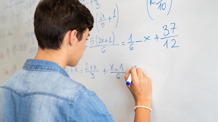 Back view of high school student solving math problem on whiteboard in classroom. Young man writing math solution on white board using marker. College guy solving math expression during lesson.