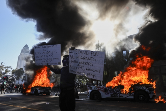 A man holds signs as a Waymo vehicles burn, as protesters clash with law enforcement in the streets surrounding the federal building during a protest following federal immigration operations in Los Angeles, California, on June 8, 2025. Demonstrators torched cars and scuffled with security forces in Los Angeles on June 8, as police kept protestors away from the National Guard troops President Donald Trump sent to the streets of the second biggest US city. (Photo by RINGO CHIU / AFP)
